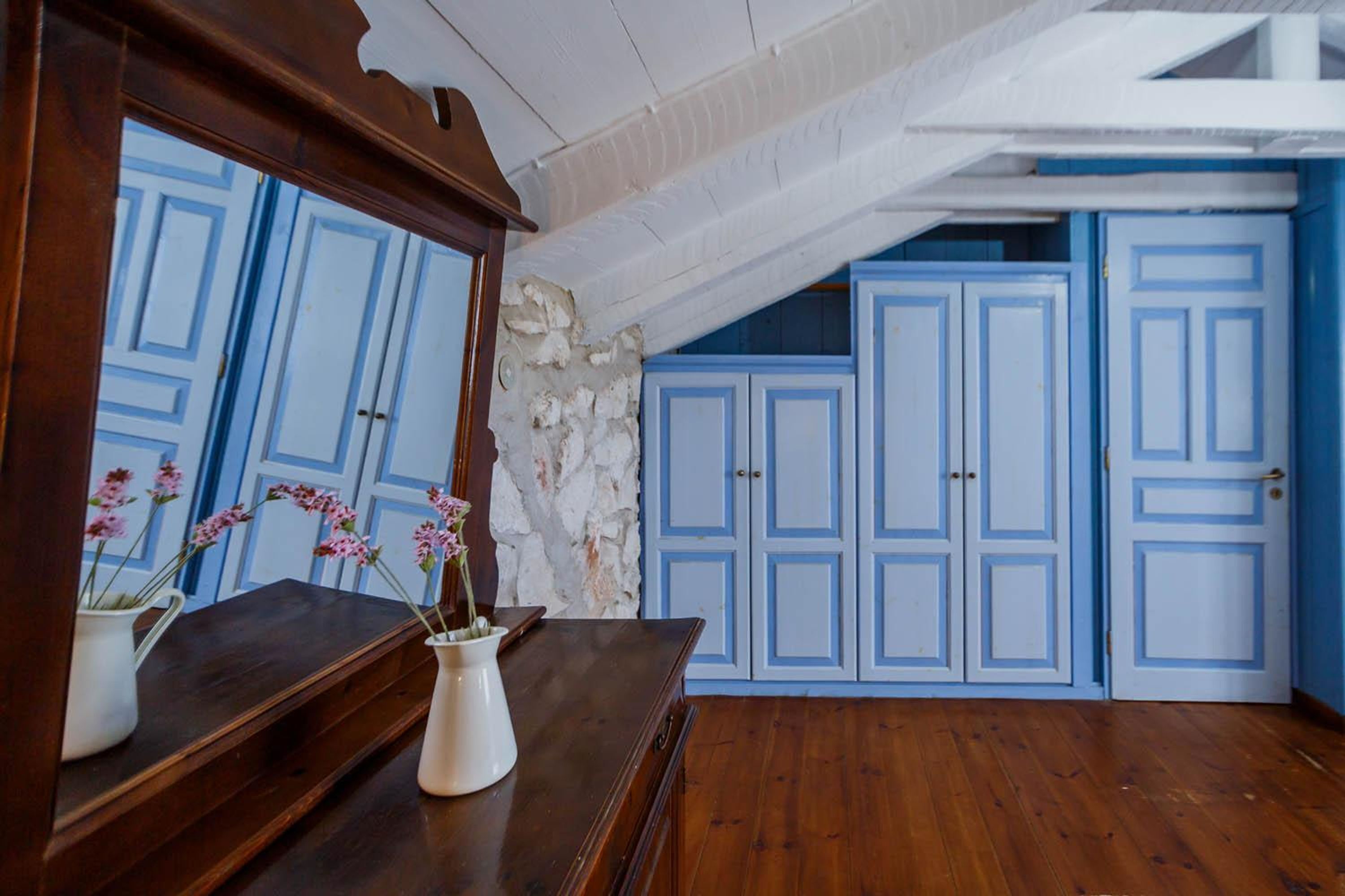 A room with a wooden dresser, a mirror, and a vase with flowers. There are blue doors and a stone wall in the background.