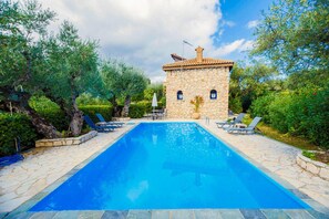 Ein Swimmingpool mit Liegestühlen, ein Steinhaus und üppiges Grün.