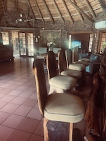 Interior rústico com telhado de palha, paredes de tijolo e um espaço de estar com cadeiras estofadas.