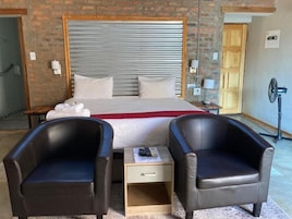Quarto de hotel com uma cama grande, duas poltronas de couro, uma mesa de cabeceira com controle remoto e um ventilador.