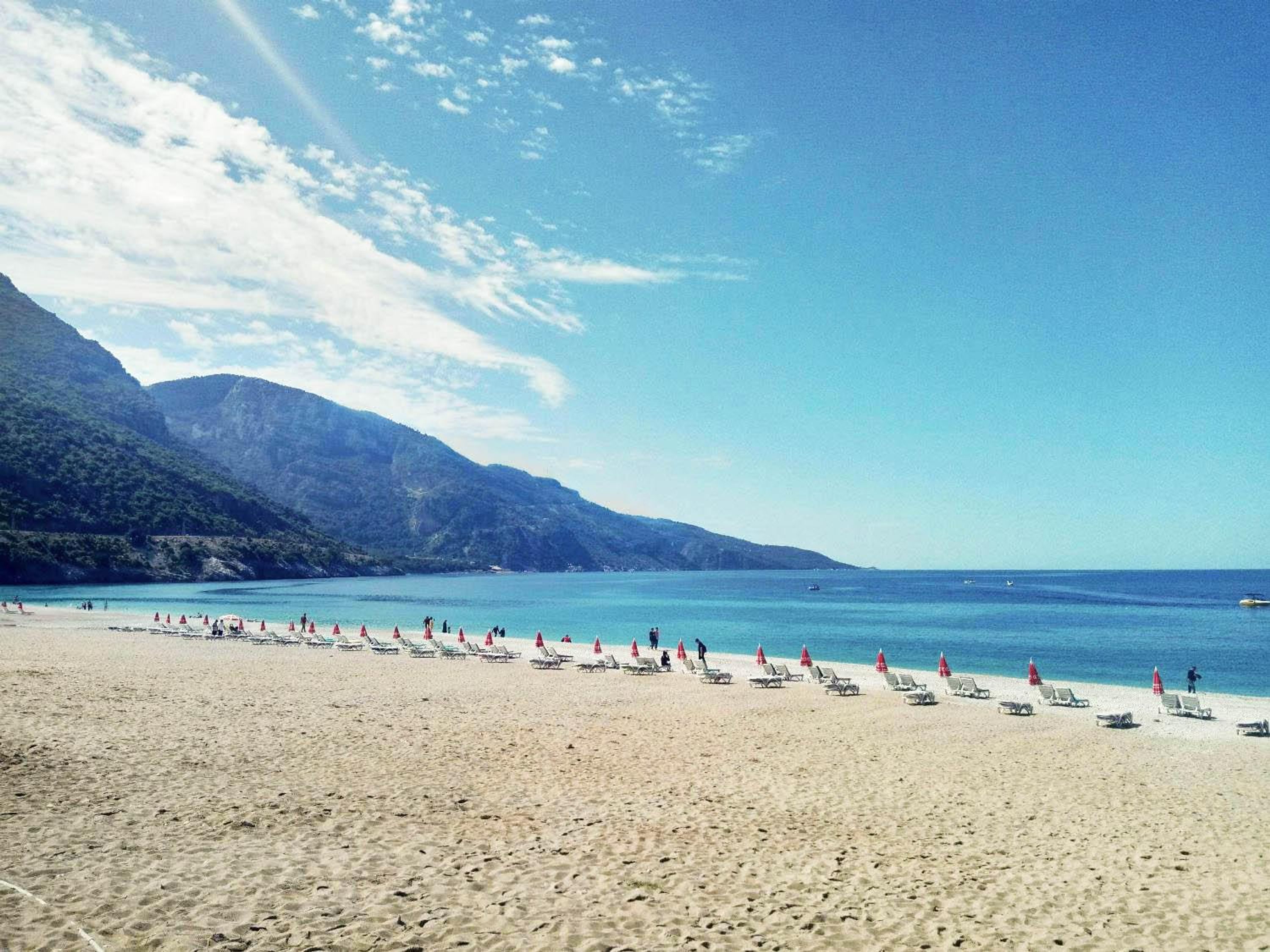 Ein Strand mit Reihen von Liegestühlen und Sonnenschirmen, einem klaren Himmel und Bergen im Hintergrund.