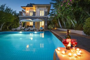 A swimming pool with lounge chairs, a table with candles and wine glasses, and a building with stone facade and balcony. - Villa Tara in Oludeniz (Fethiye)