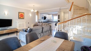 A modern living room with a sofa, a dining table, and a TV. A kitchen is visible in the background, and a staircase leads to another level. - Villa Tara in Oludeniz (Fethiye)