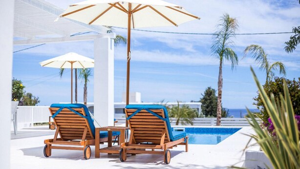 Ein Poolbereich mit zwei Liegestühlen und einem Tisch, einem großen Sonnenschirm und Blick aufs Meer.