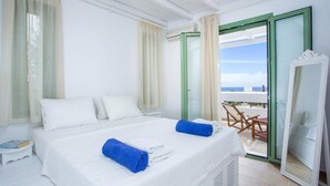 Ein Hotelzimmer mit einem Bett, zwei blauen Handtüchern, einem weißen Nachttisch, einem Sessel und Blick auf das Meer durch eine Glastür.