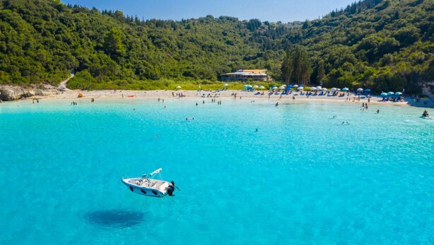 A beach with clear turquoise water, a boat, and a building in the background. - Villa Magda Dione in Gaios (Paxos)