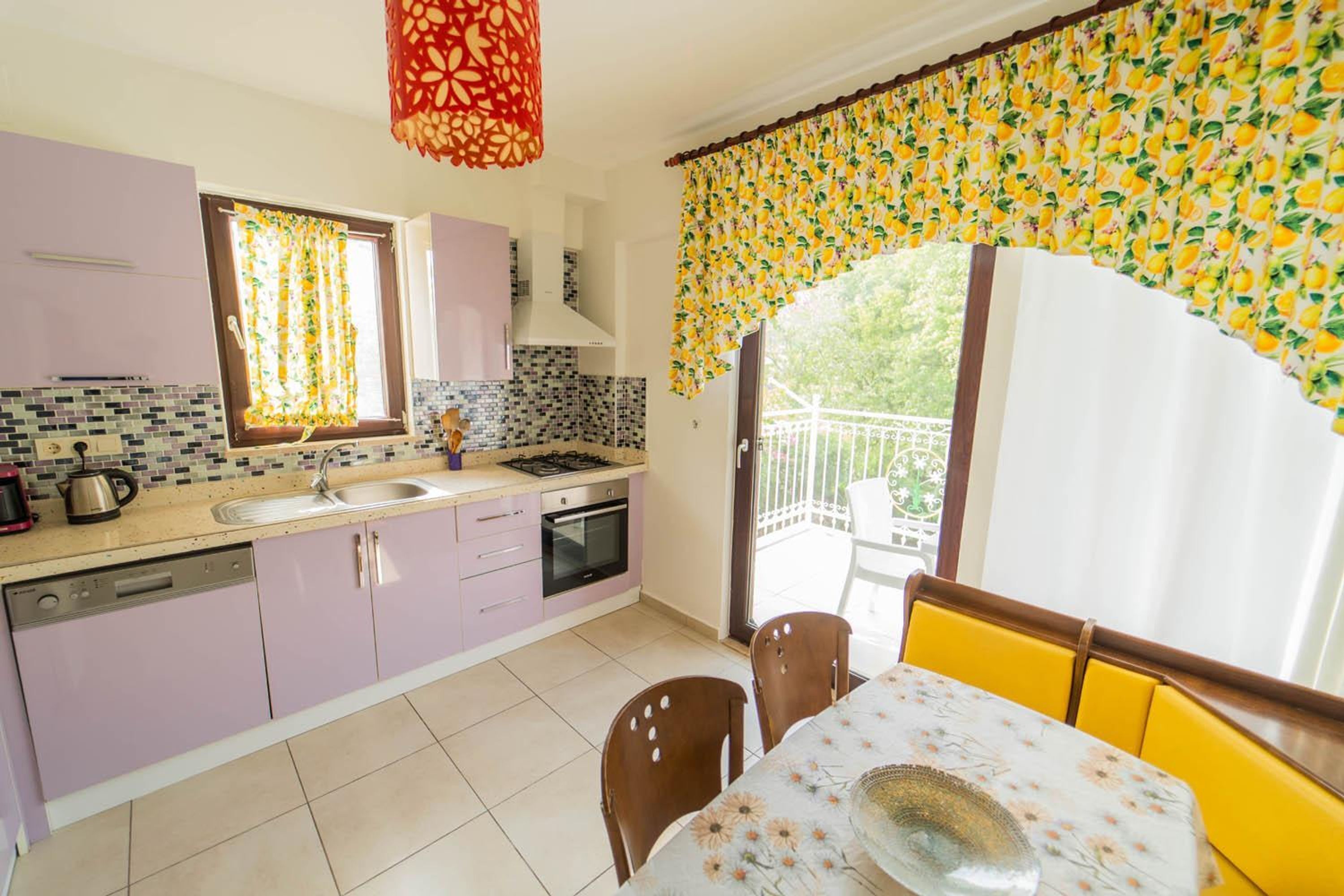 Una cocina con lavamanos, estufa y horno, una mesa de comedor con sillas, y un balcón con vistas al verdor.