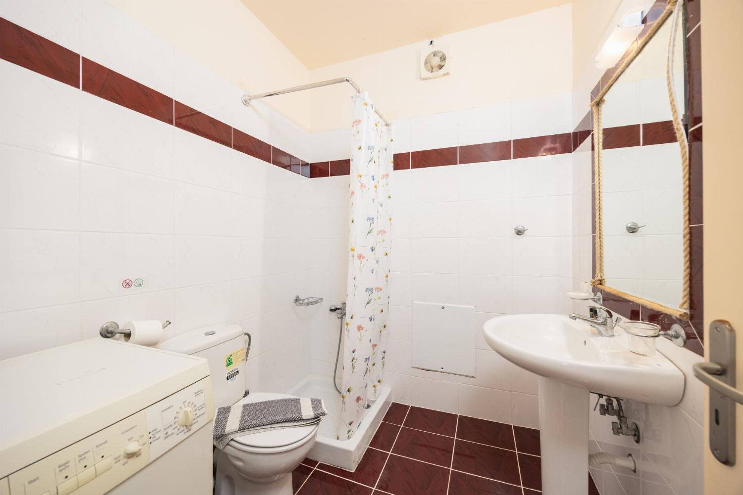 Uma casa de banho com sanita, máquina de lavar roupa, chuveiro com cortina floral, banheira e lavatório com espelho.