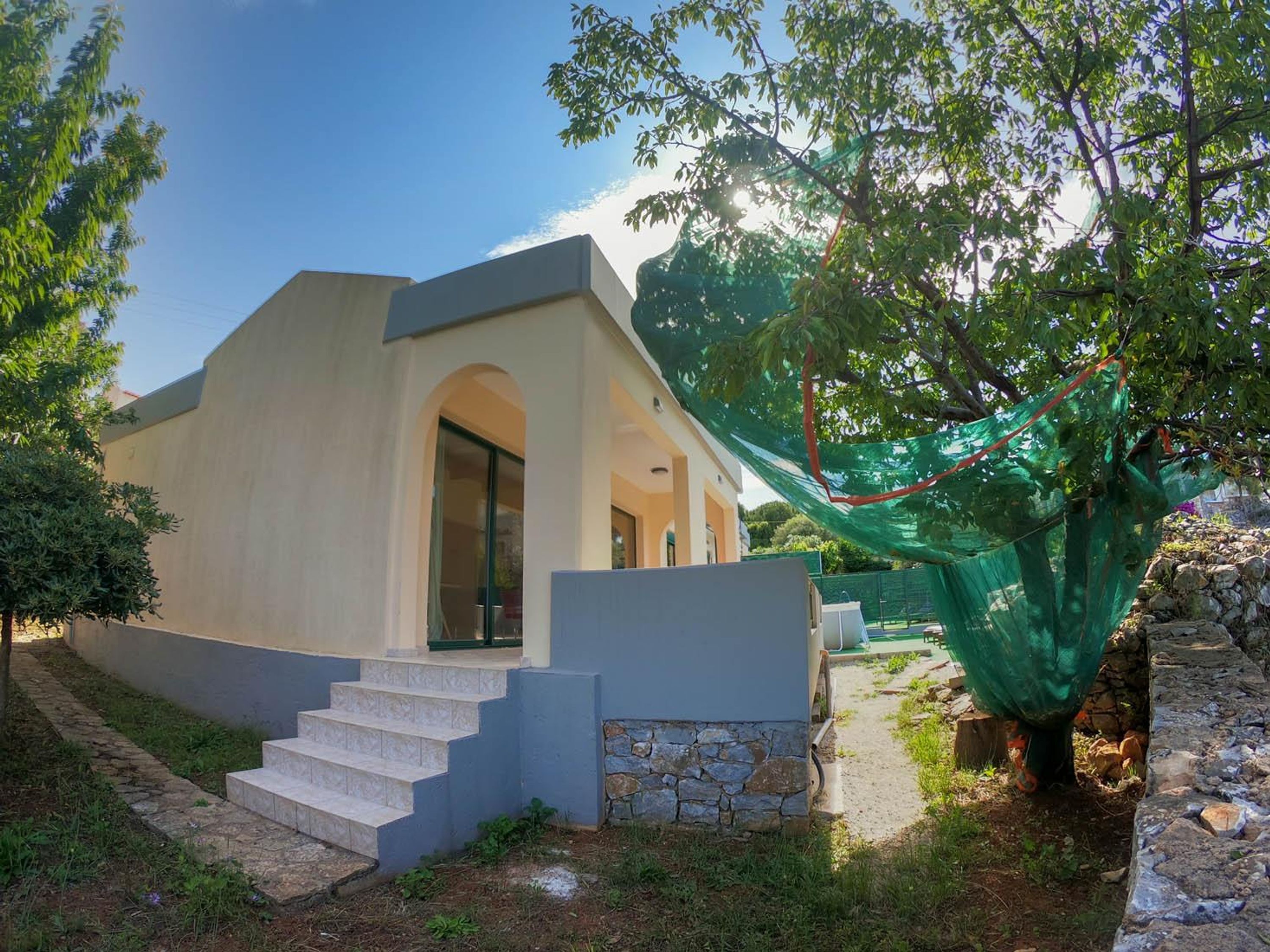 A modern house with a stone foundation, a set of stairs leading to an entrance, and a tree with a green netting.