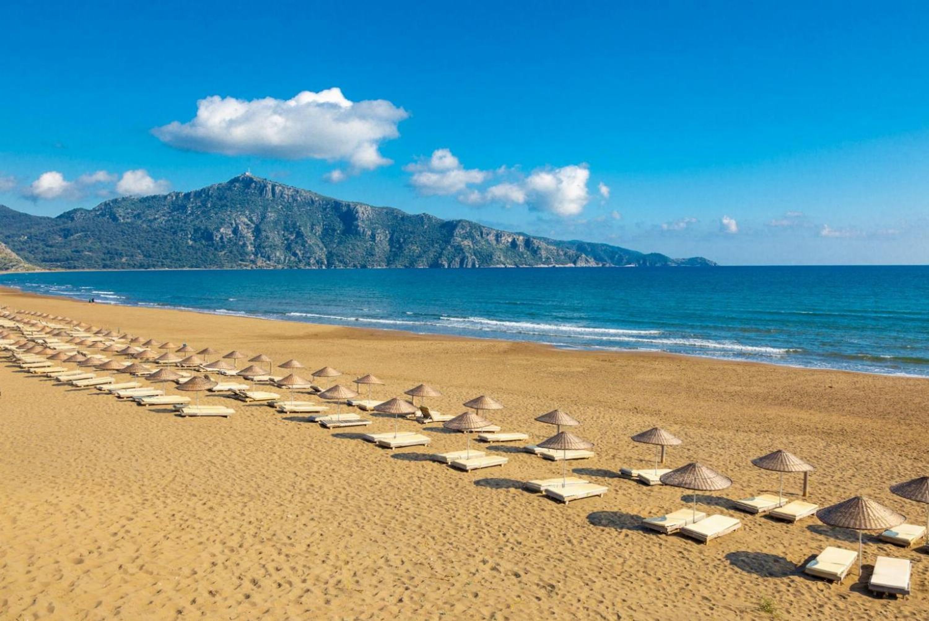 A beach with rows of sun loungers and umbrellas, a clear blue sky, and a mountain in the background.