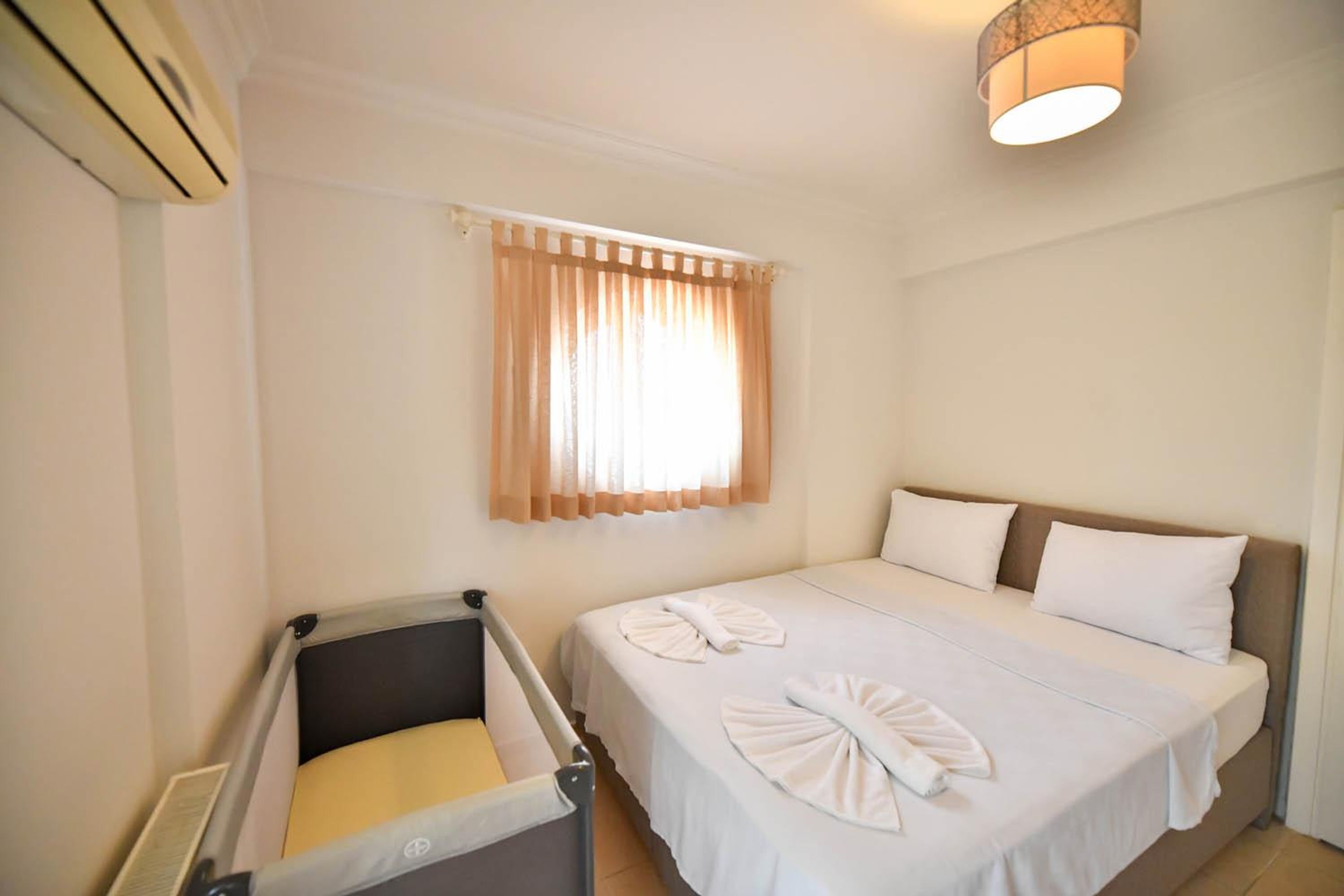 A hotel room with a bed, a small bedside table, a window with curtains, and an air conditioning unit.