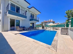 Ett modernt hus i två våningar med en swimmingpool, en uteplats och ett grönt staket.