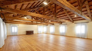 Ein geräumiger Raum mit Holzboden, einer hohen Decke mit sichtbaren Holzbalken und großen Fenstern mit weißen Vorhängen.