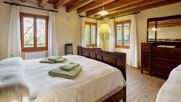 Een slaapkamer met een houten plafond, een bed met een witte dekbedovertrek, twee ramen met gordijnen, een bedlamp en een houten dressoir.