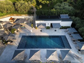 Vista aérea de uma área de piscina moderna ao ar livre, com uma piscina retangular, cercada por espreguiçadeiras e guarda-sóis, e um edifício com telhado plano.
