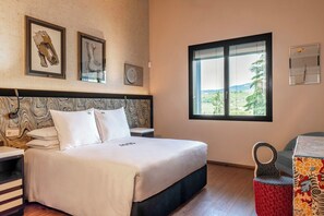 Quarto de hotel com uma cama, mesa de cabeceira, uma cadeira, janela com vista e duas obras de arte emolduradas na parede.