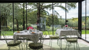 Un comedor con mesas dispuestas para cuatro personas, rodeado de sillas de metal con asientos acolchados, y ventanas amplias que dan a un exuberante jardín.