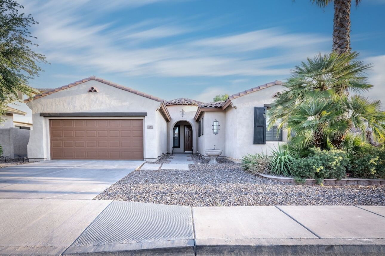 A two-story house with a brown garage door, a front entrance with a door, and a small garden with palm trees and shrubs.
