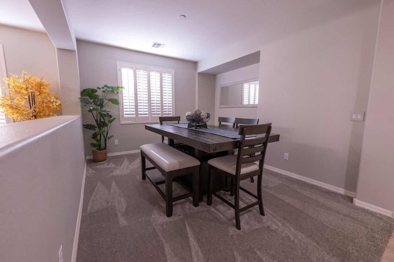 A dining area with a dark wooden table, four chairs, and a bench. There are two windows with blinds, a potted plant, and a vase with flowers on the table.