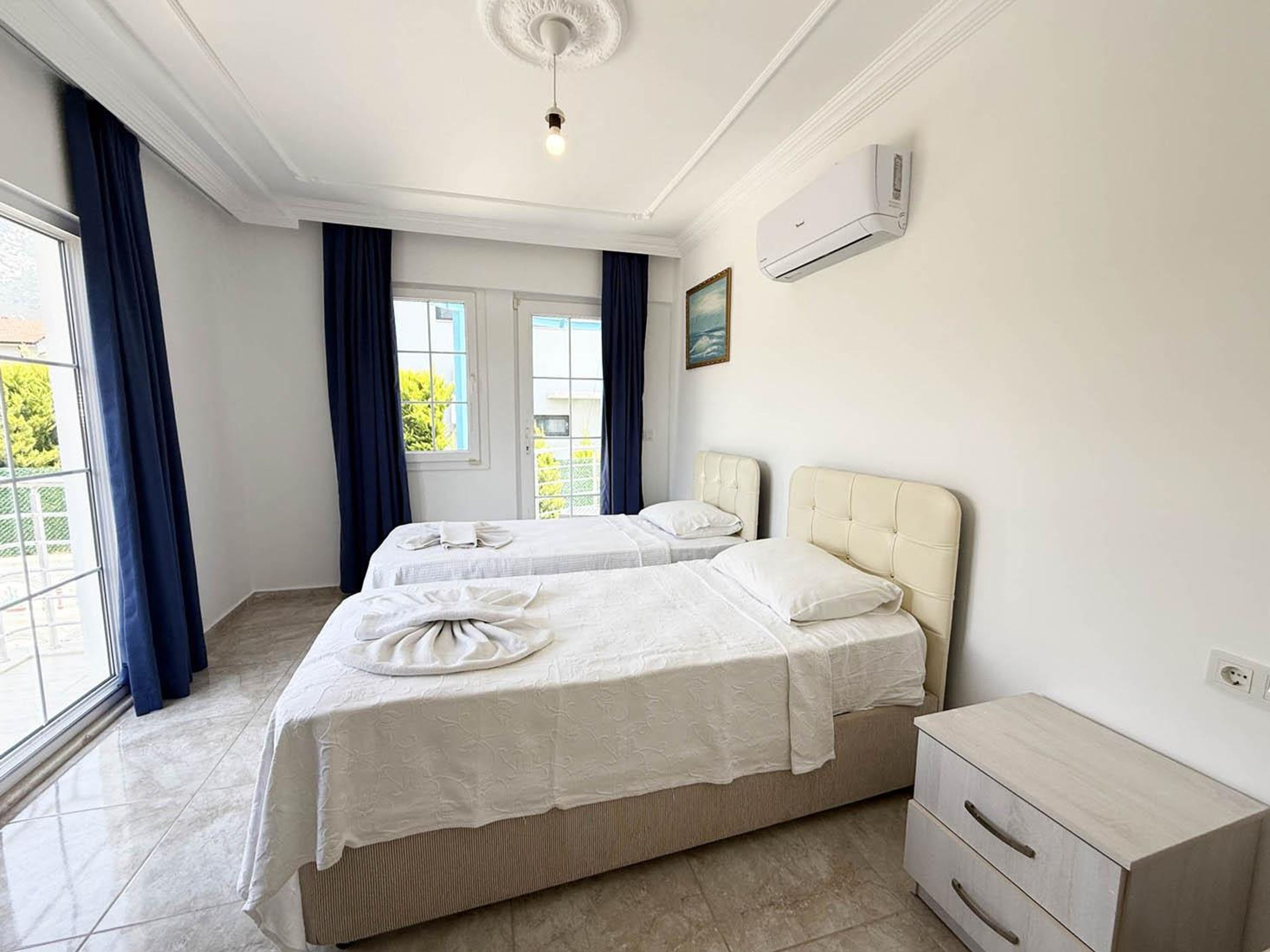 A hotel room with two beds, a window with curtains, a painting on the wall, and an air conditioning unit.