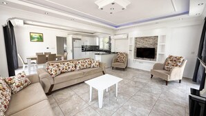 A spacious living room with a sofa, chairs, a coffee table, and a TV. There is a kitchen area in the background.