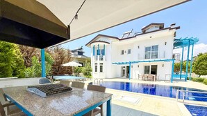 An outdoor pool area with a covered seating area, a pool table, and a two-story house in the background.