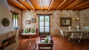 A rustic stone-walled room with wooden beams, a stone fireplace, a wooden sofa, a dining table with chairs, and a large window with green shutters. - Villa Faidra in Agios Dimitrios (Zakynthos)