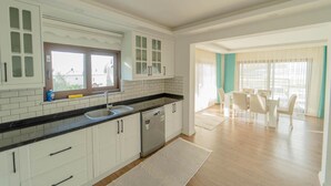 A modern kitchen with white cabinets, a black countertop, and a built-in dishwasher. There is a dining area with a table and chairs, and a large window providing natural light. - Villa Everest in Oludeniz (Dalaman)