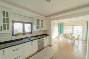 A modern kitchen with white cabinets, a black countertop, and a built-in dishwasher. There is a dining area with a table and chairs, and a large window providing natural light. - Villa Everest in Oludeniz (Dalaman)