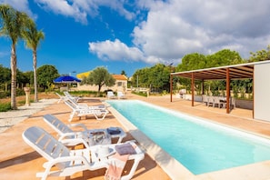 A swimming pool with lounge chairs and umbrellas, surrounded by trees and a building with a red roof. - Villa Fenice in Scicli (Scicli)