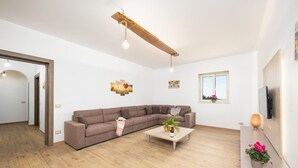 Een moderne woonkamer met een bruine hoekbank, een houten salontafel met planten, een houten balk in het plafond, en een aan de muur bevestigde televisie.