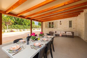 An outdoor dining area with a long table set for four, wooden beams overhead, stone walls, and a view of greenery. - Villa Fenice in Scicli (Scicli)