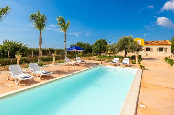 A swimming pool with lounge chairs and umbrellas, surrounded by palm trees and a building with a yellow roof. - Villa Fenice in Scicli (Scicli)