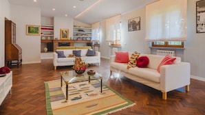Un salon spacieux avec un canapé blanc, une table basse en verre et un tapis à motifs. On y trouve une cheminée avec un manteau de cheminée, des étagères et un escalier menant à un autre niveau.