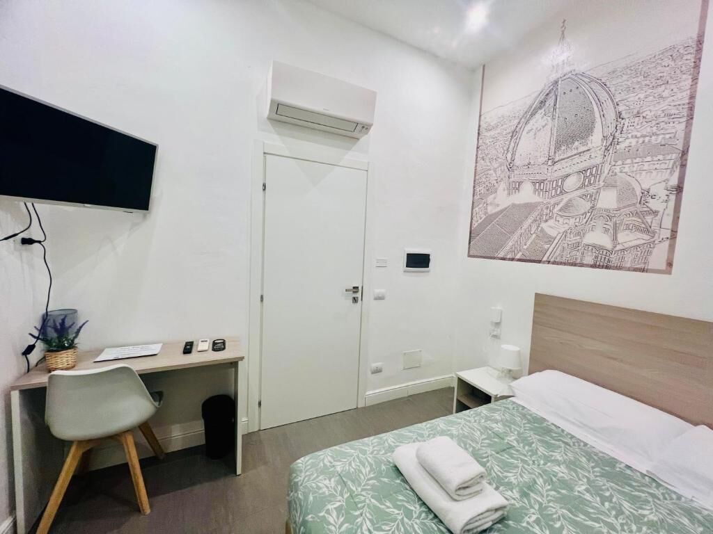 A hotel room with a bed, a desk with a chair, a flat-screen TV, an air conditioner, and a wall-mounted artwork.