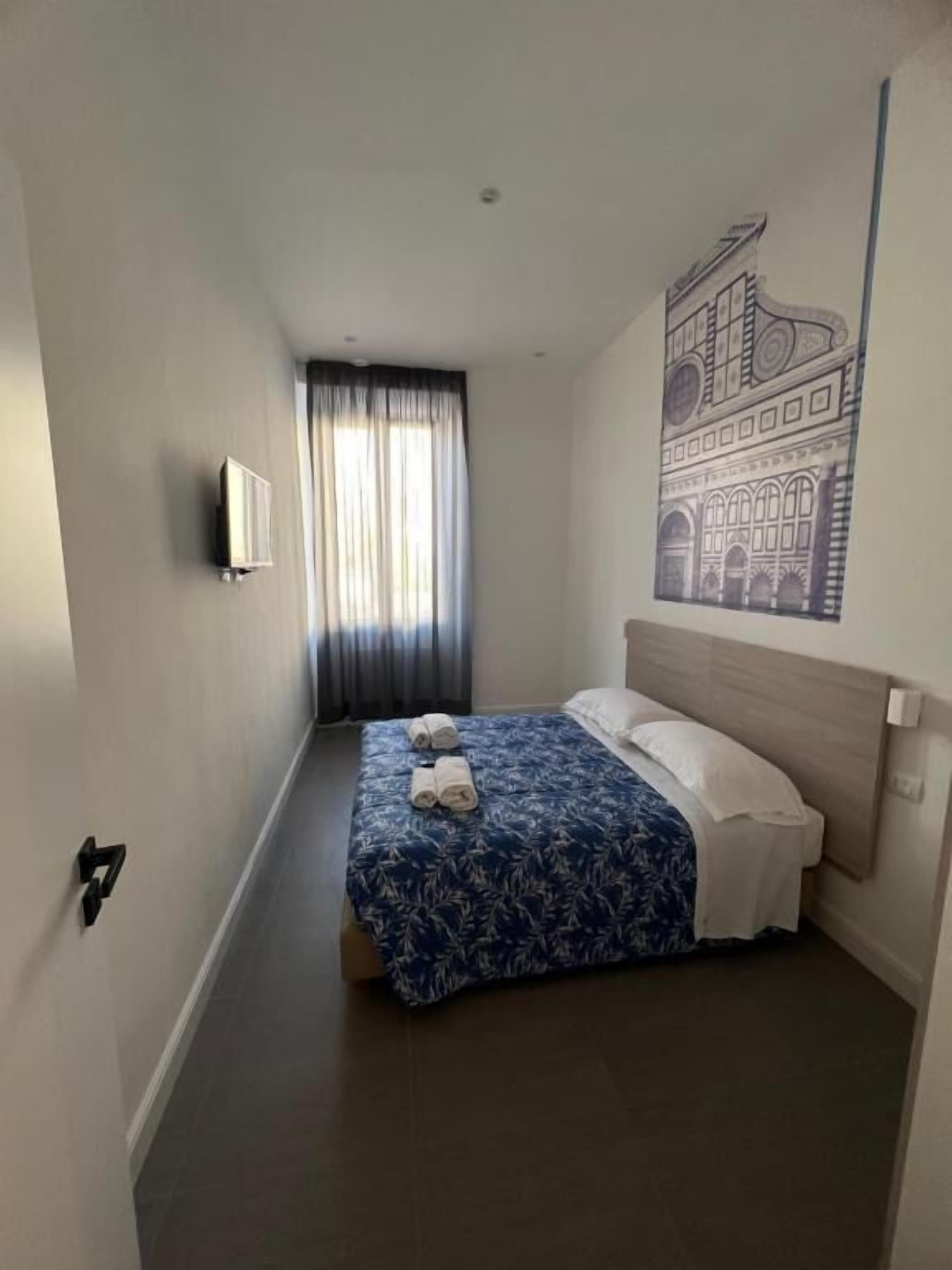 Quarto de hotel com cama, televisão fixada na parede, janela com cortinas e um quadro na parede.