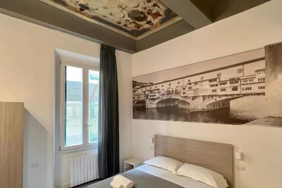 A hotel room with a bed, a nightstand, a window with curtains, and a large cityscape painting on the wall.