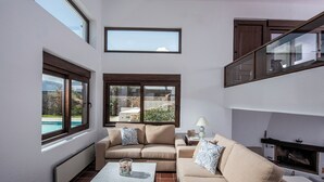 Ein Wohnzimmer mit zwei Sofas, einem Couchtisch und einem Kamin. Große Fenster bieten Ausblick ins Freie.