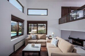 Ein Wohnzimmer mit zwei Sofas, einem Couchtisch und einem Kamin. Große Fenster bieten Ausblick ins Freie.