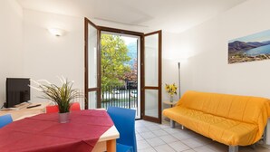 A room with a dining table, chairs, a sofa, a TV, and a plant. - Mobilehome in Porlezza am Luganersee (Porlezza)