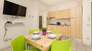 A compact kitchen with wooden cabinets, a flat-screen TV mounted on the wall, and a dining table set for four with green chairs. - Mobilehome in Porlezza am Luganersee (Porlezza)
