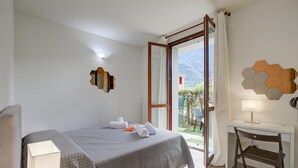 A hotel room with a bed, a desk, a chair, a window with a view of mountains, and a door leading to an outdoor area. - Mobilehome in Porlezza am Luganersee (Porlezza)