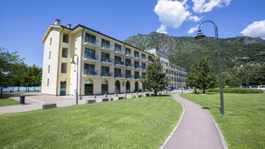 A multi-story hotel with a yellow facade, a paved walkway, and a mountainous backdrop. - Mobilehome in Porlezza am Luganersee (Porlezza)