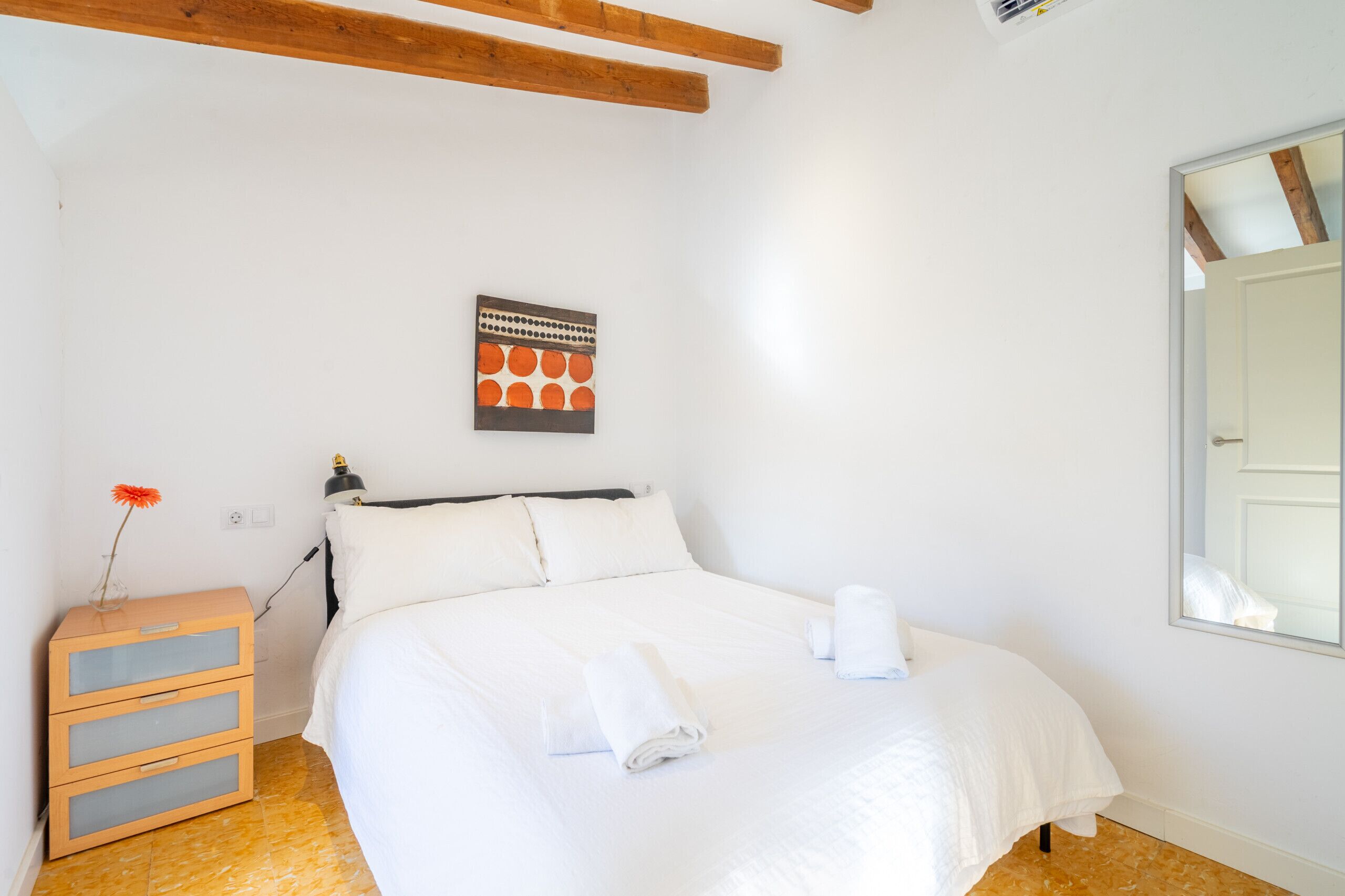 A neatly made bed with white linens, a wooden nightstand with drawers, a framed artwork on the wall, and a large window with a view of the sky.