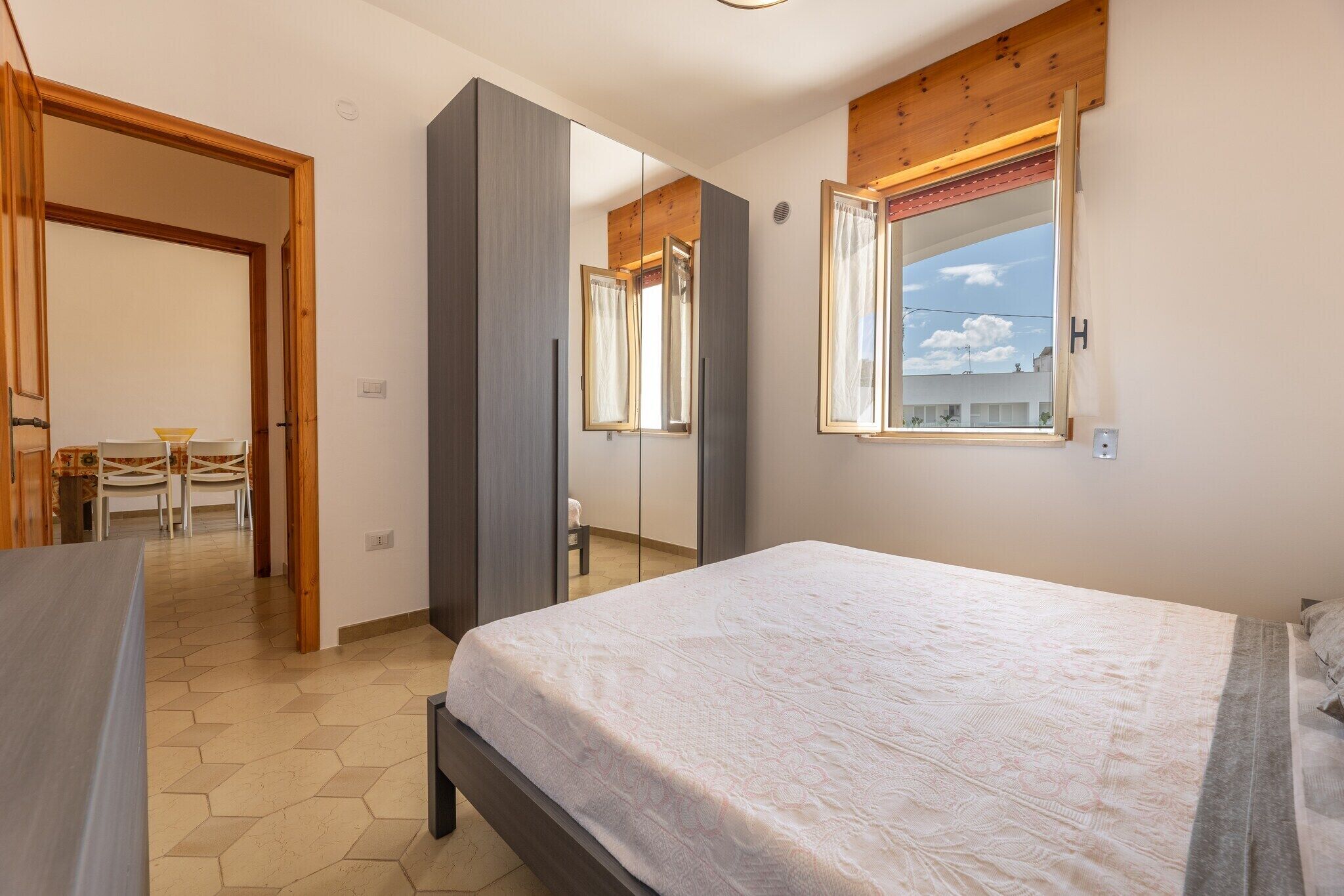 A bedroom with a large bed, a wardrobe, a mirror, and a window with a view of the sky.