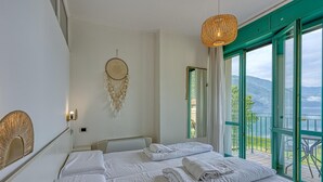 A hotel room with a bed, a dreamcatcher, a hanging light, and a balcony with a view of the mountains. - Appartement mit Seeblick auf den Luganersee (Porlezza)
