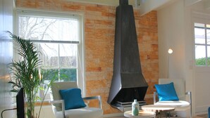 Uma sala de estar moderna com uma parede de pedra, uma lareira, duas cadeiras brancas com almofadas azuis, uma mesa de centro redonda e uma planta grande.