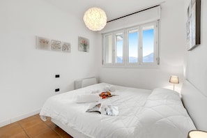 Una cama bien hecha con edredón blanco, una lámpara de noche y una ventana con vistas al cielo y a las montañas.