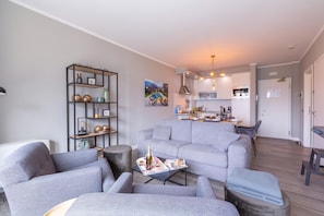 Una sala de estar moderna con un sofá gris, una mesa de centro con revistas y una estantería. Se ve una cocina al fondo, con armarios blancos y una lámpara colgante.