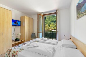 A hotel room with two beds, a TV, a desk with a plant, and a balcony with a view of greenery. - Mobilehome in Porlezza am Luganersee (Porlezza)